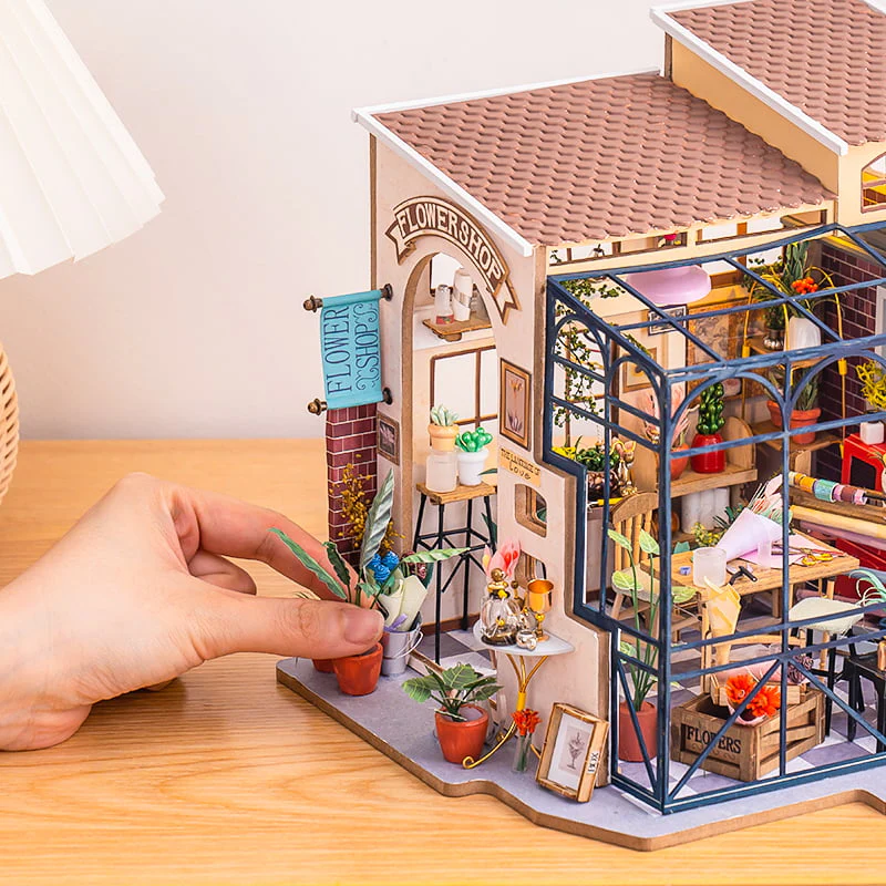 A hand gently placing a potted plant on the fully assembled Rolife Emily's Flower Shop Miniature Kit resting on a table.