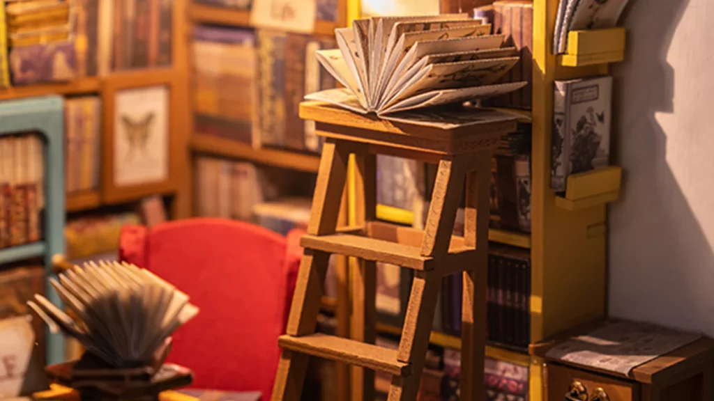 A flipped-over book resting on a short wooden ladder and another on a table with a red chair in the Rolife Sam's Study Library Kit.