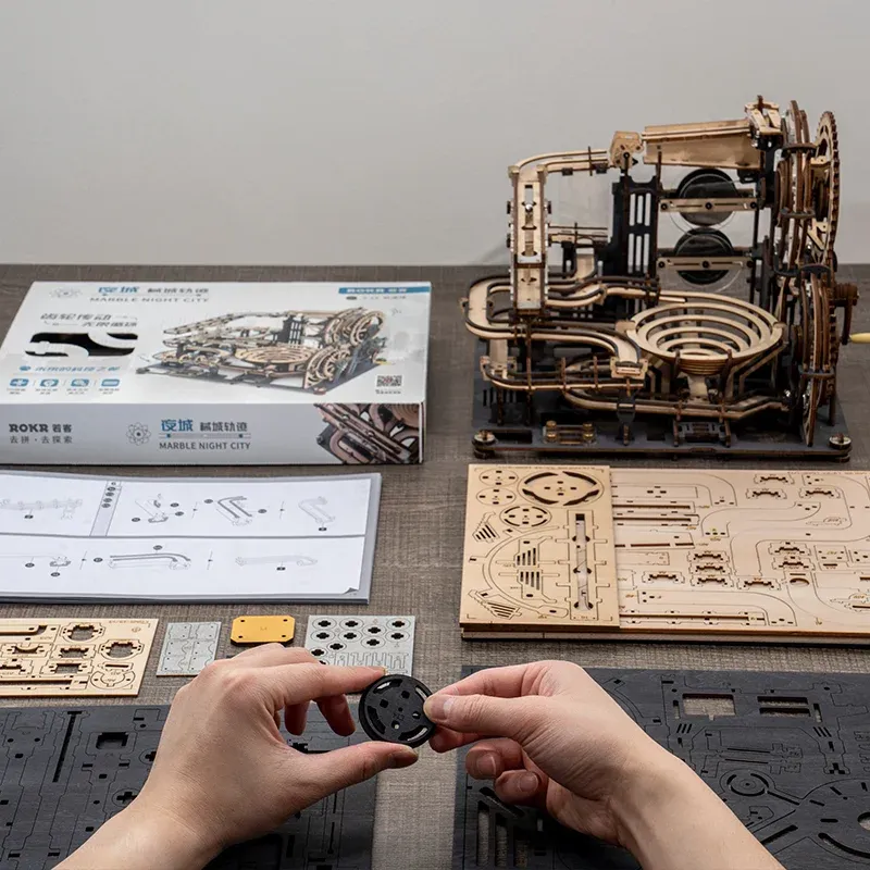 Hands assembling Marble Night City Marble Run 3D puzzle kit on a dark wood table, with fully assembled puzzle, templates, and box neatly arranged.