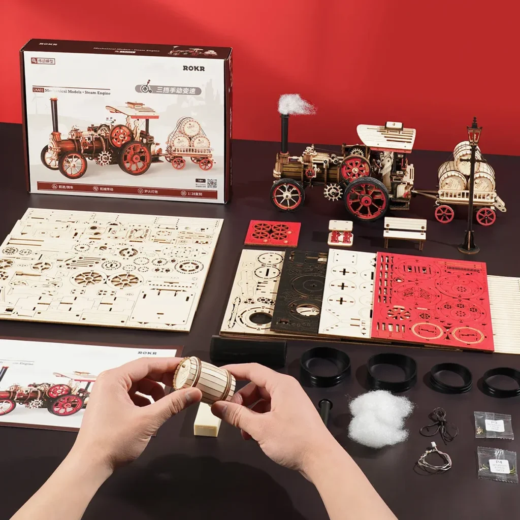 Hands assembling the steam engine tractor 3D puzzle kit on a table, featuring the box, templates, and fully constructed model.