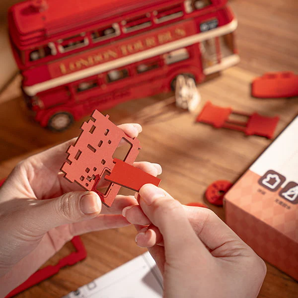 Hands assembling the Rolife London Tour Bus 3D Wooden Puzzle kit on a wooden table, with a partially completed bus, precut parts, and packaging.