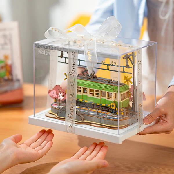 Hands exchanging the Sakura Journey 3D Wooden Puzzle in a clear display case with a white ribbon.
