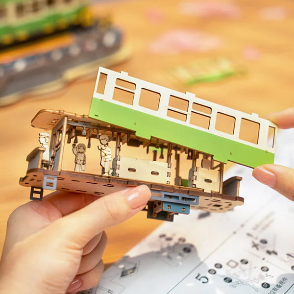 Hands assembling the Sakura Journey 3D Wooden Puzzle tram car on a wooden table.